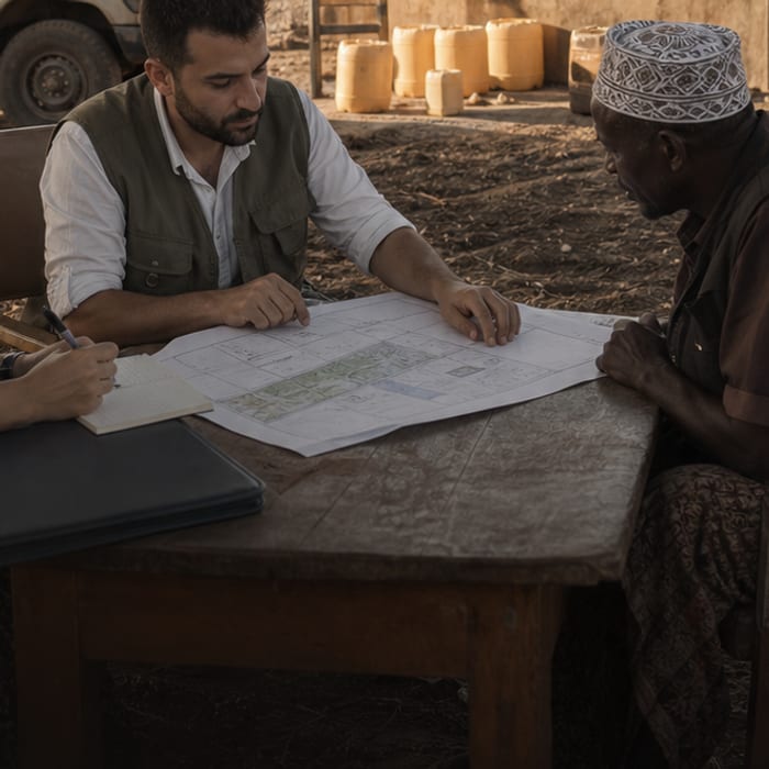 Saha masasının üzerinde açılmış proje planı; Vuslat Organizasyon görevlisi ve yerel temsilci planın üzerinde ayrıntıları gösteriyor.